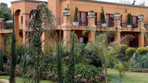 A large, multi-story building with a symmetrical facade, balconies with ornate railings, and a well-maintained garden with various shrubs and palm trees. - Tuareg Guest House (Midrand)