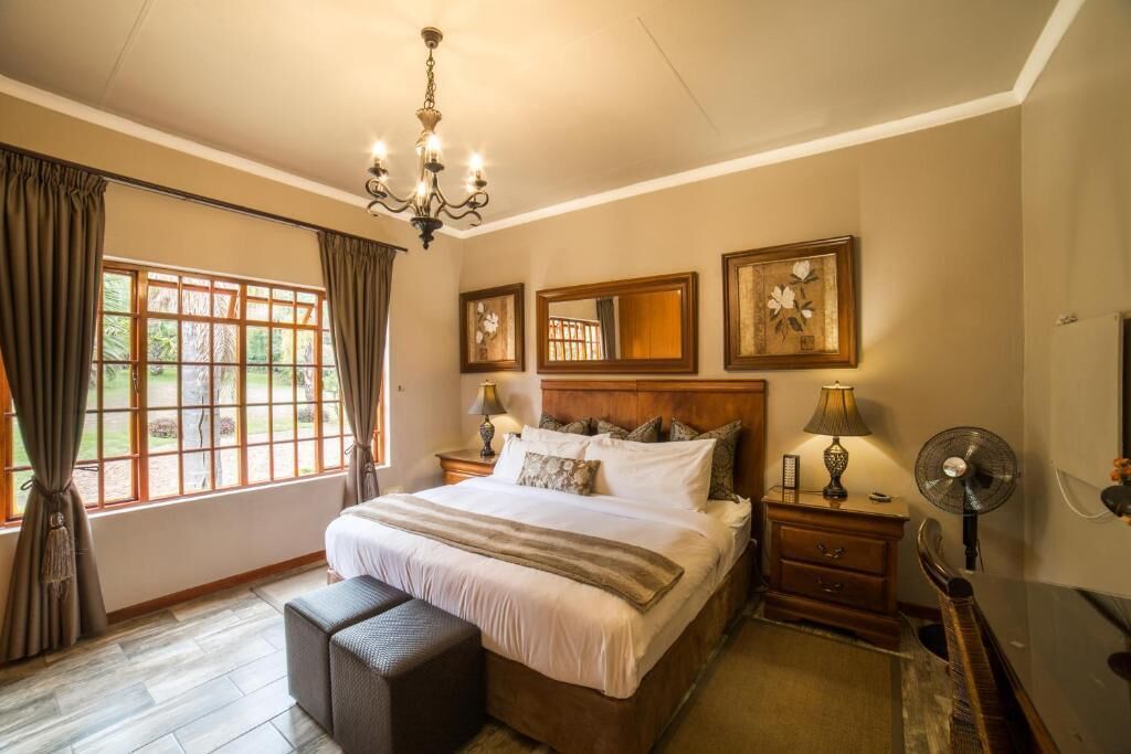 A hotel room with a large bed, a chandelier, a window with curtains, a bedside table with a lamp, and a mirror on the wall.