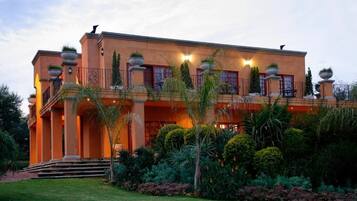 Um edifício grande, de vários andares, com fachada simétrica, várias varandas e um jardim bem cuidado com diversas plantas.