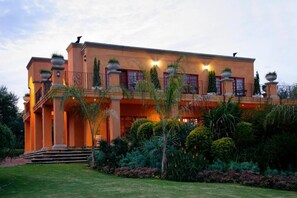 Un edificio grande de varios pisos con una fachada simétrica, múltiples balcones y un jardín bien cuidado con diversas plantas.