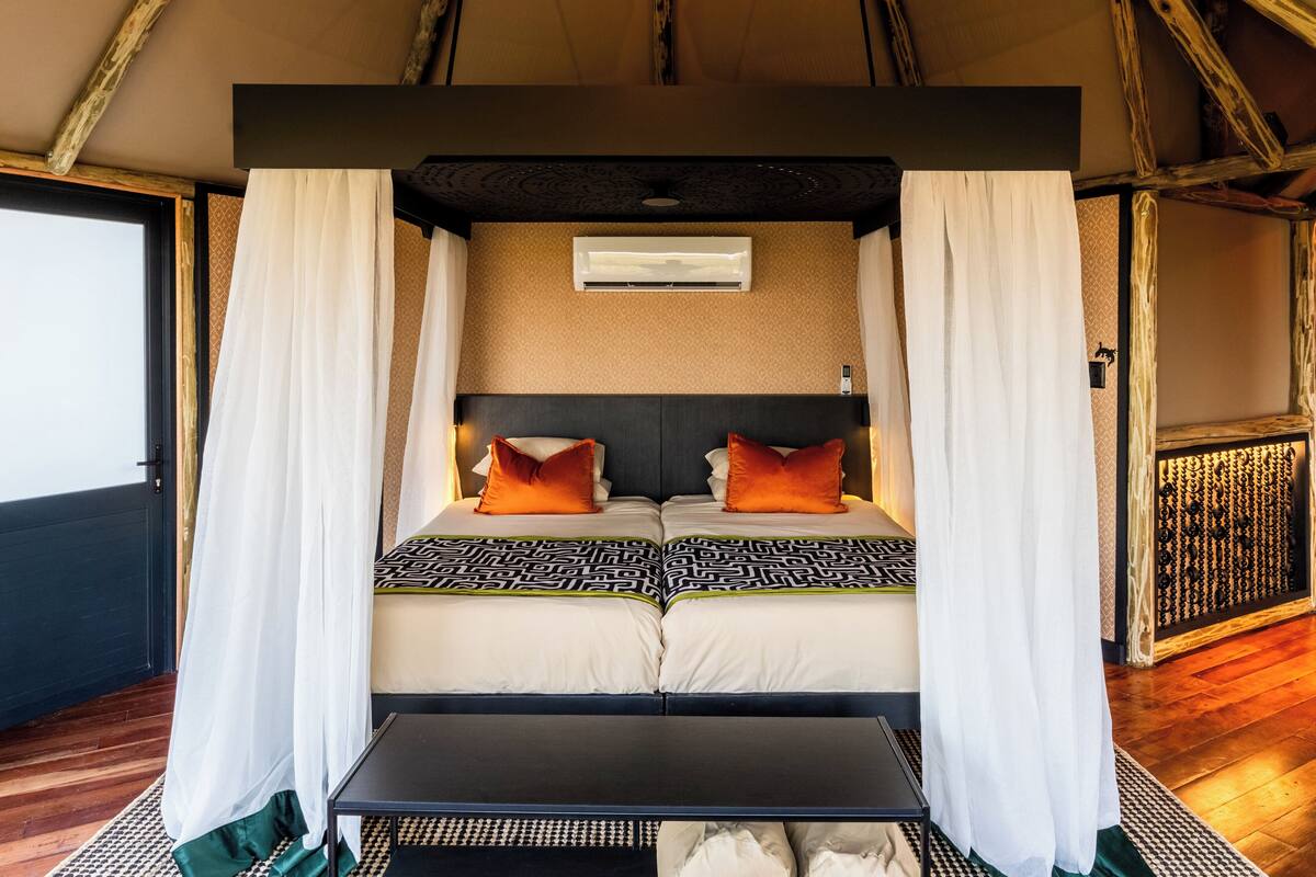 A room with a bed, white curtains, a headboard, a small table, and a patterned rug.