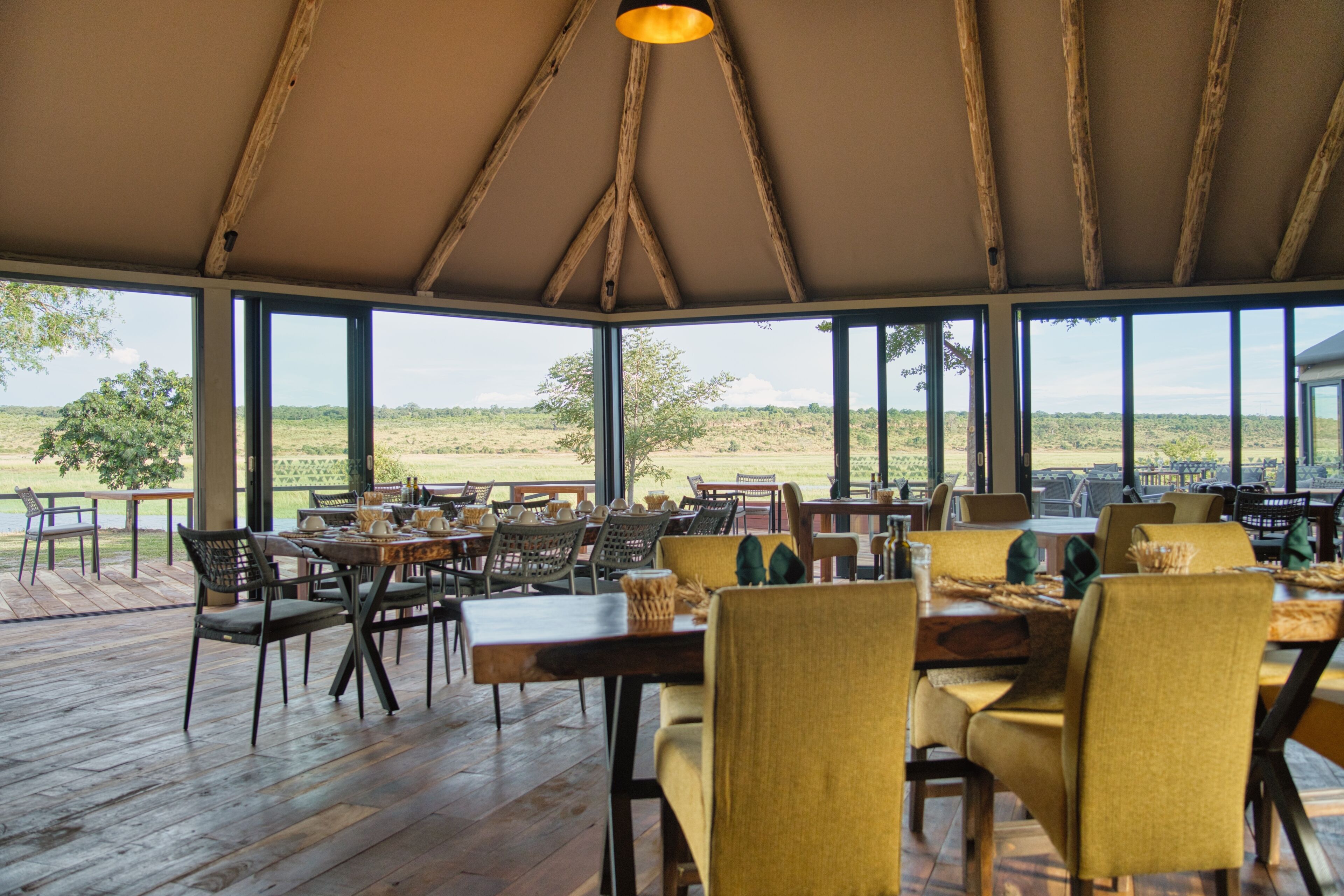 An indoor dining area with wooden tables and chairs, large windows offering a view of the outdoors.