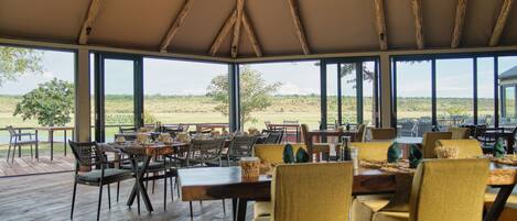 An indoor dining area with wooden tables and chairs, large windows offering a view of the outdoors.