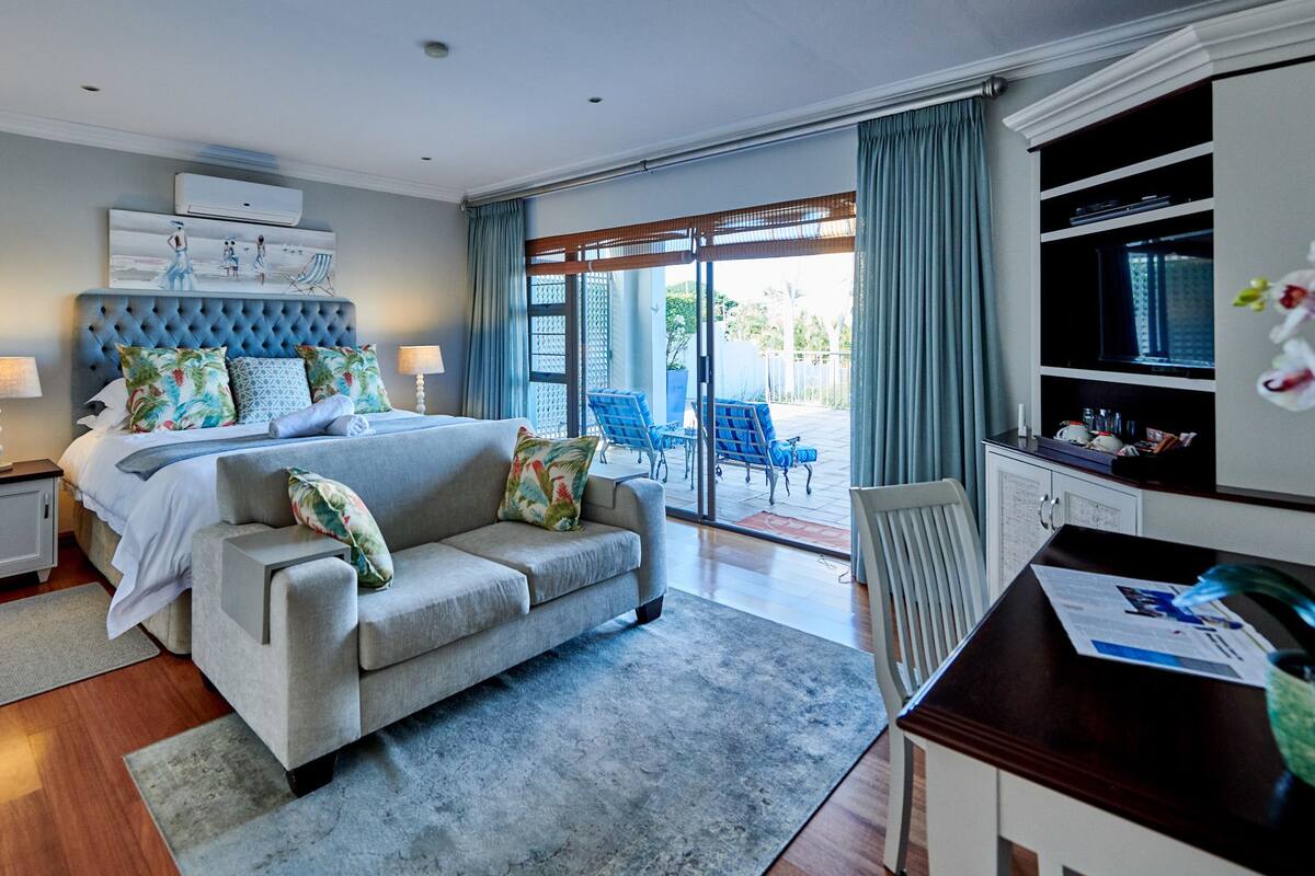 A modern bedroom with a large bed, a sofa, a desk, and a balcony with chairs.