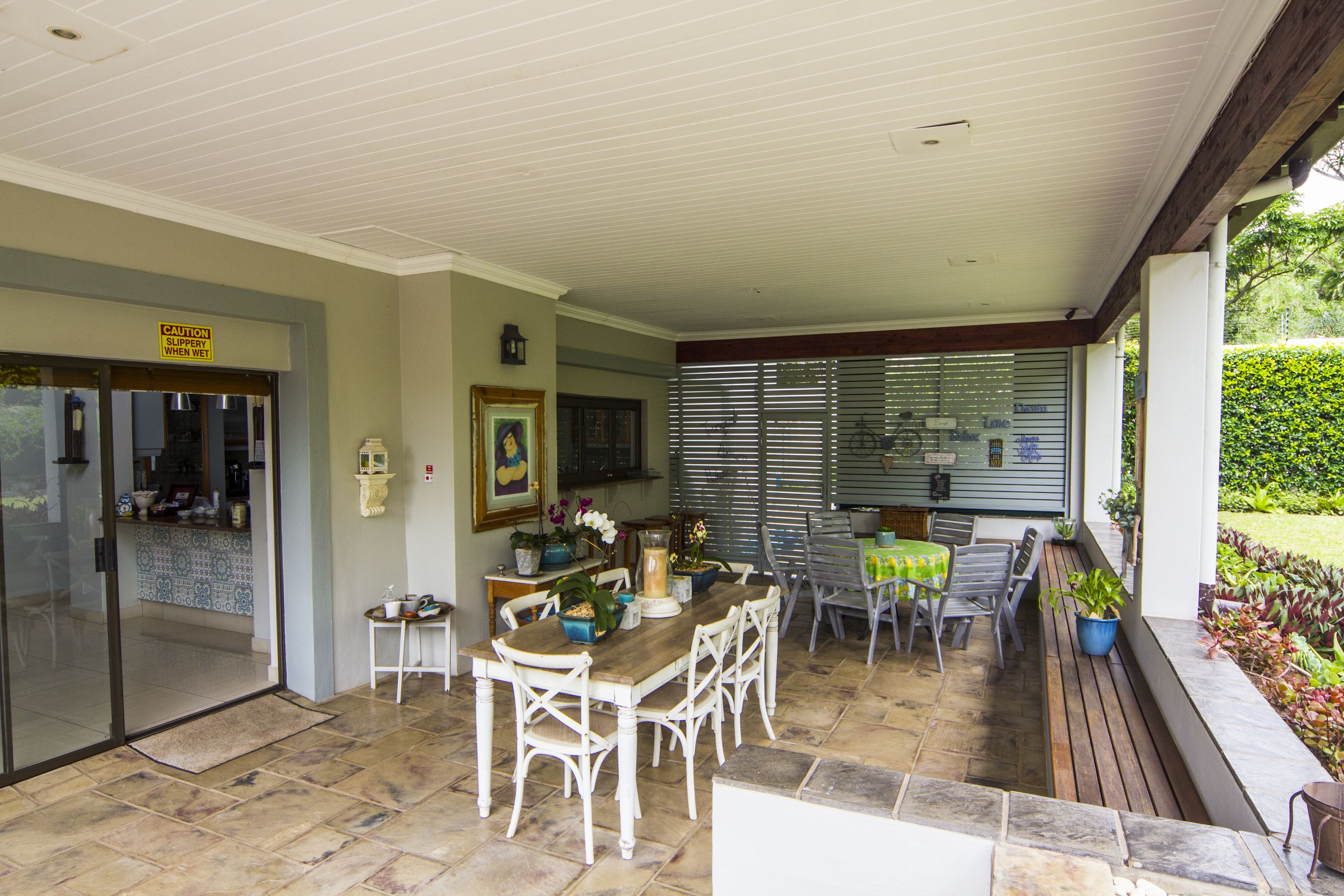 Un patio techado con una mesa de comedor y sillas, una mesita con un florero y un cartel que advierte sobre piso resbaladizo cuando está mojado.