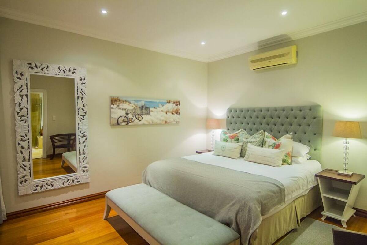 A bedroom with a large bed, a bench, a decorative mirror, a painting, and a bedside table with a lamp.