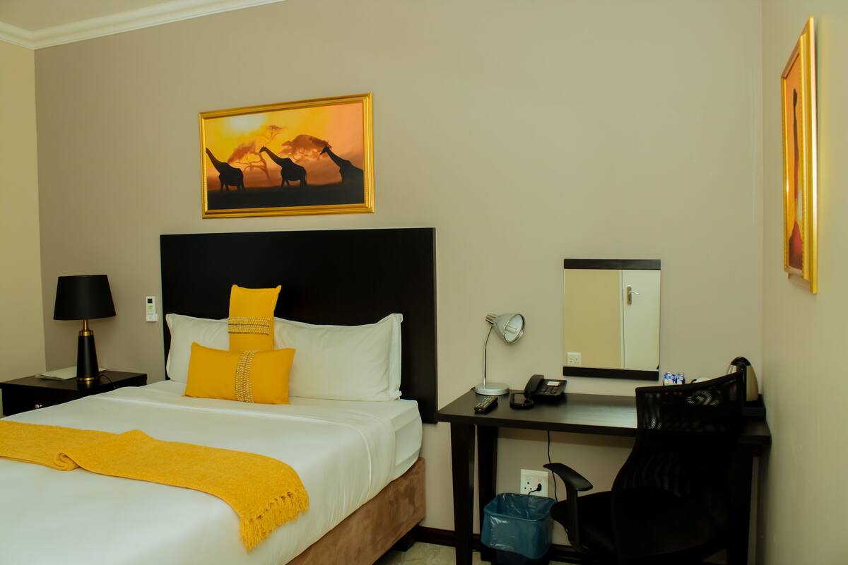A hotel room with a bed, a nightstand with a lamp, a desk with a chair, a mirror, and a painting on the wall.