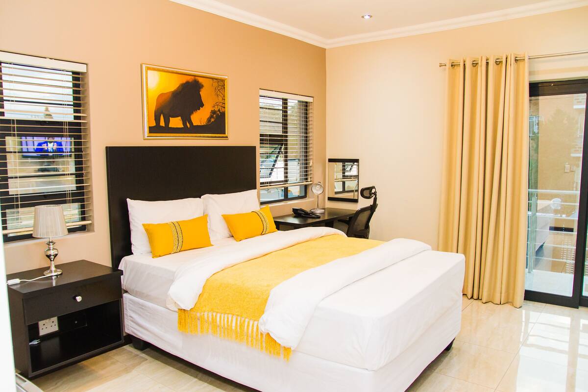 A hotel room with a large bed, a desk with a chair, a nightstand, a lamp, and a window with blinds.