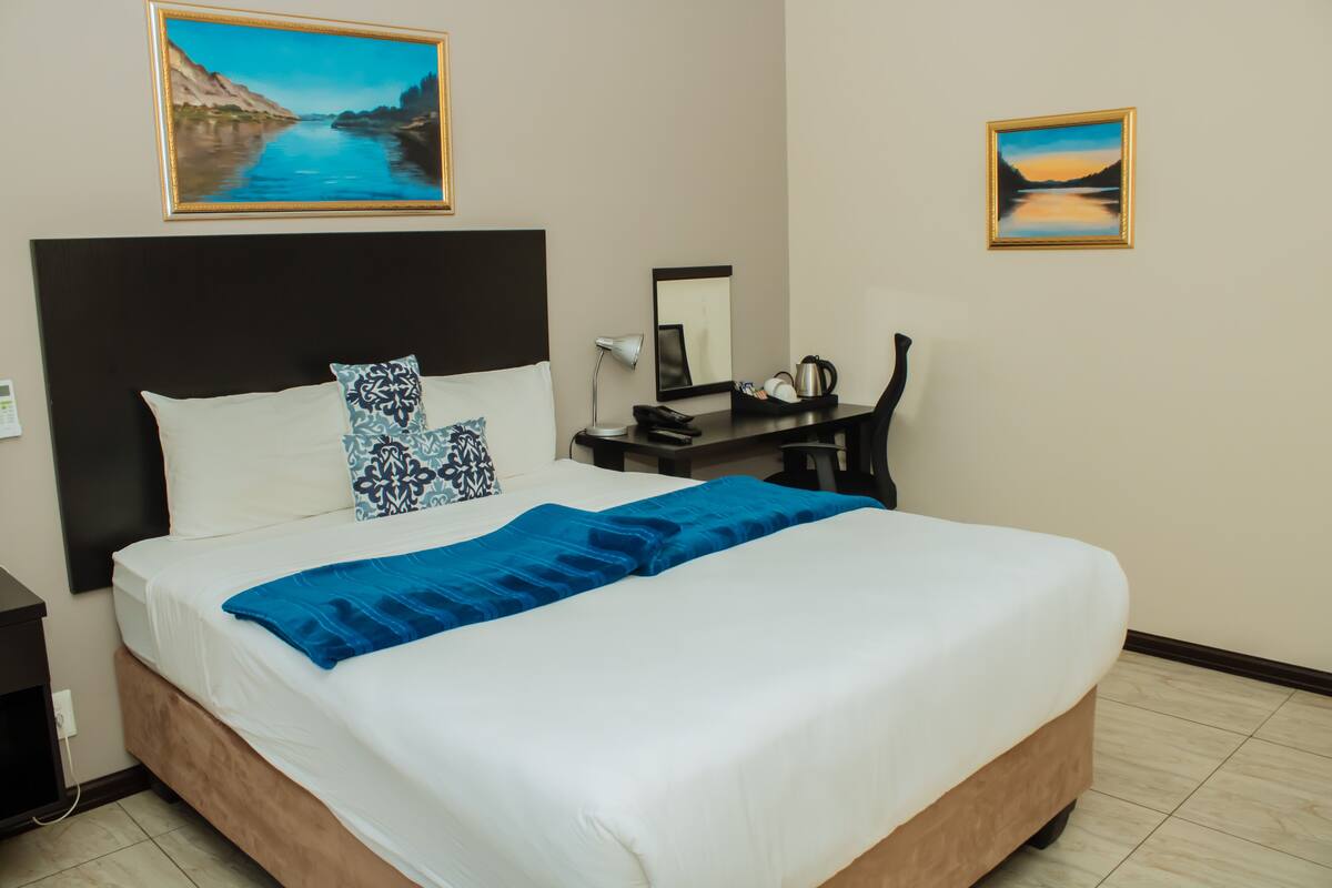 A neatly made bed with a blue blanket, a black headboard, and two patterned pillows. There is a nightstand with a lamp, a small table with a coffee pot, and a framed picture on the wall.