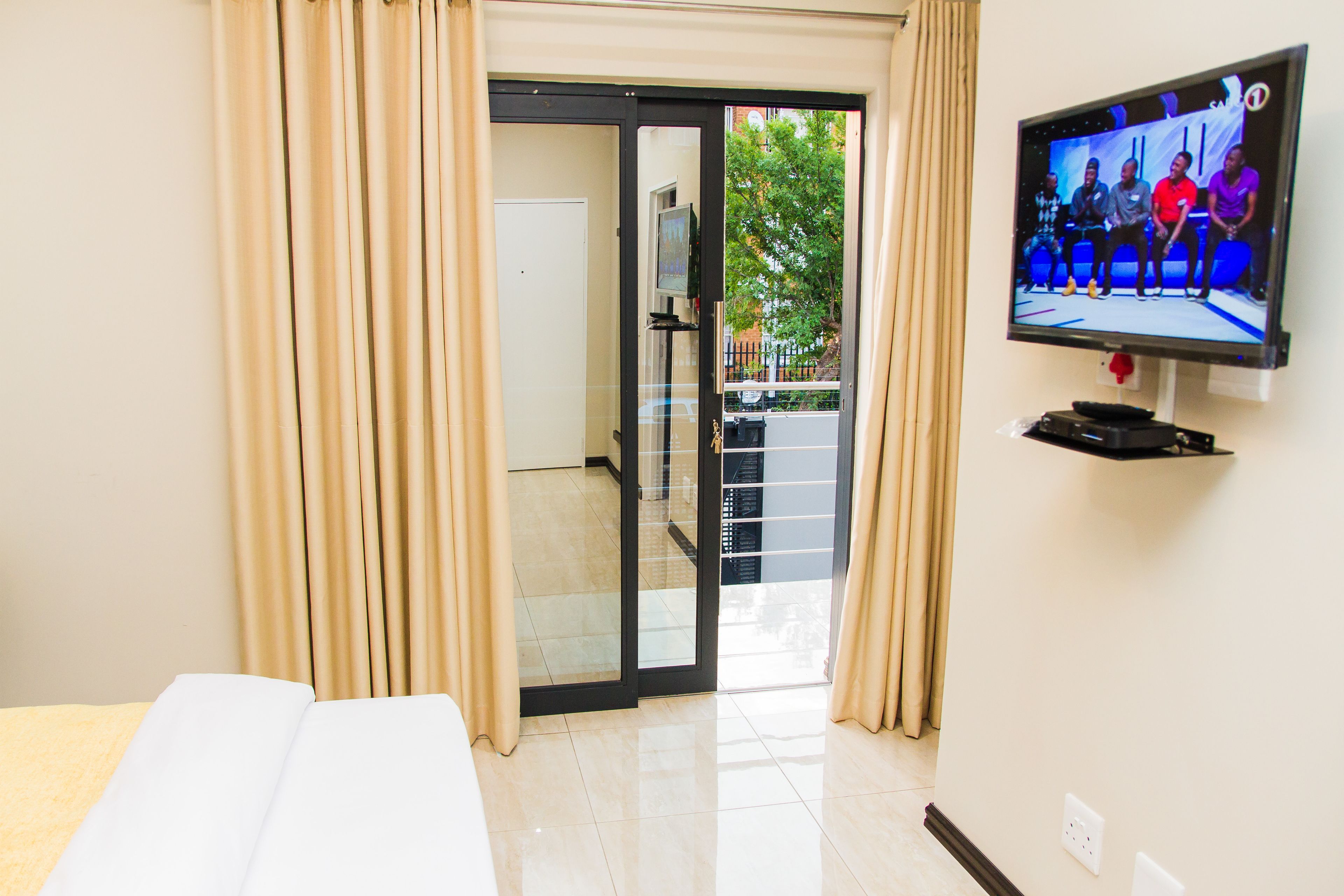 A hotel room with a sliding glass door leading to a balcony, a flat-screen TV mounted on the wall, and a bed with a white cover.