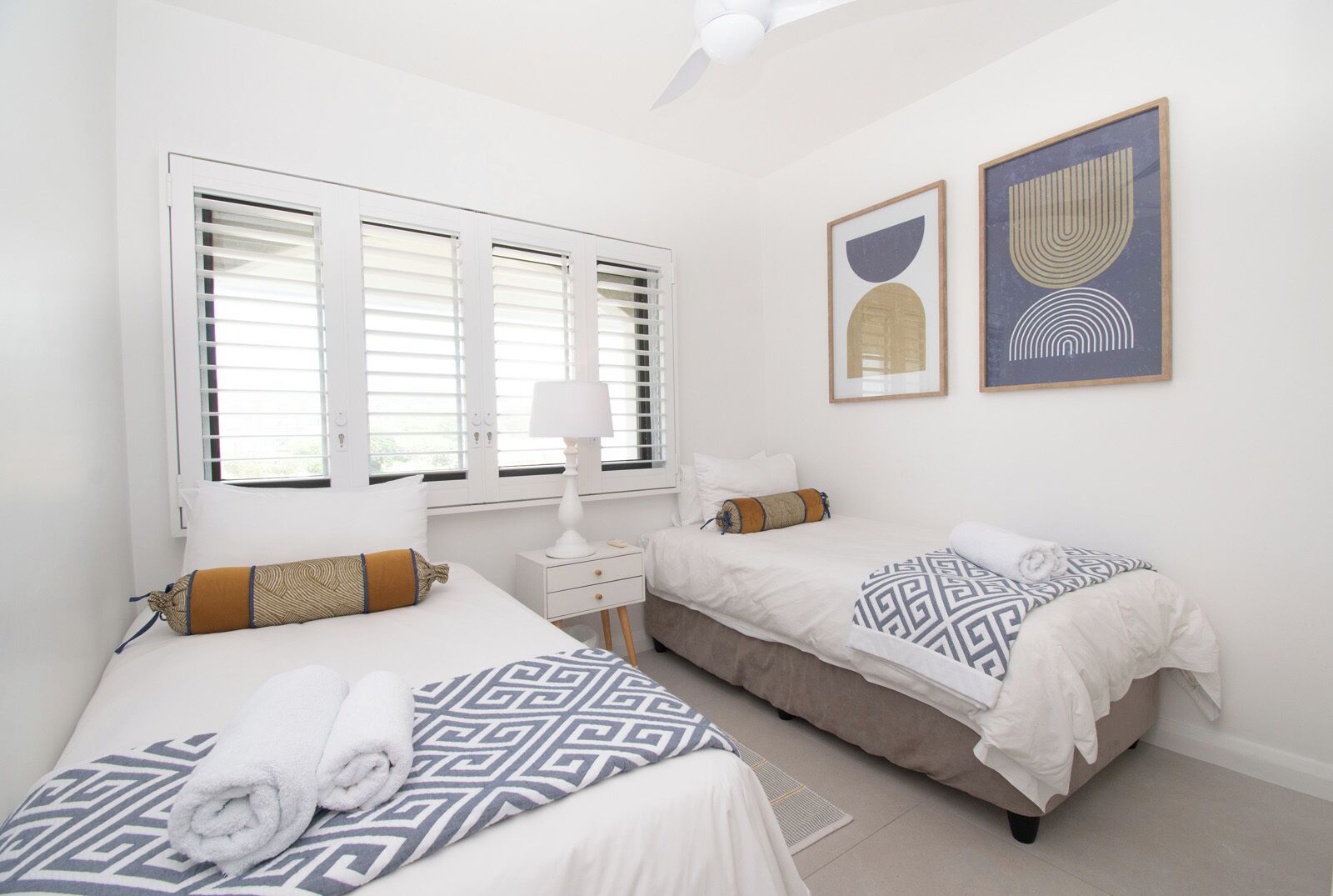A hotel room with two beds, a bedside table, a lamp, and artwork on the wall.