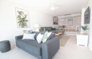 Uma sala de estar moderna com um sofá cinzento, uma mesa de centro e um espaço de jantar com cadeiras brancas.