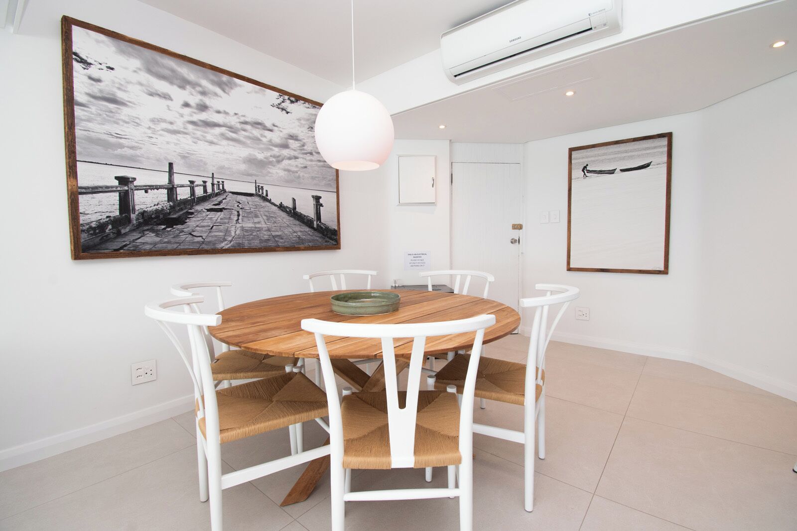 A dining area with a round wooden table, white chairs, a hanging pendant light, and a large framed photograph on the wall.