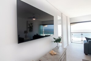 A modern living room with a large flat-screen TV mounted on the wall, a ceiling fan, a potted plant, and a view of the ocean through large windows. - 1002 Bermudas (Umhlanga)