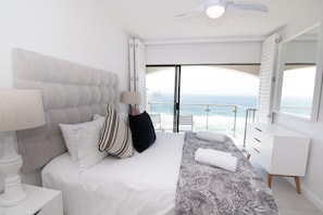 A bedroom with a bed, bedside table, lamp, and a view of the ocean.