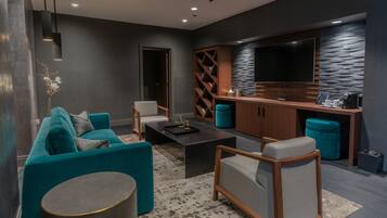 A modern living room with a sofa, coffee table, and ottoman. There is a television mounted on the wall and a small kitchen area with a countertop and appliances.