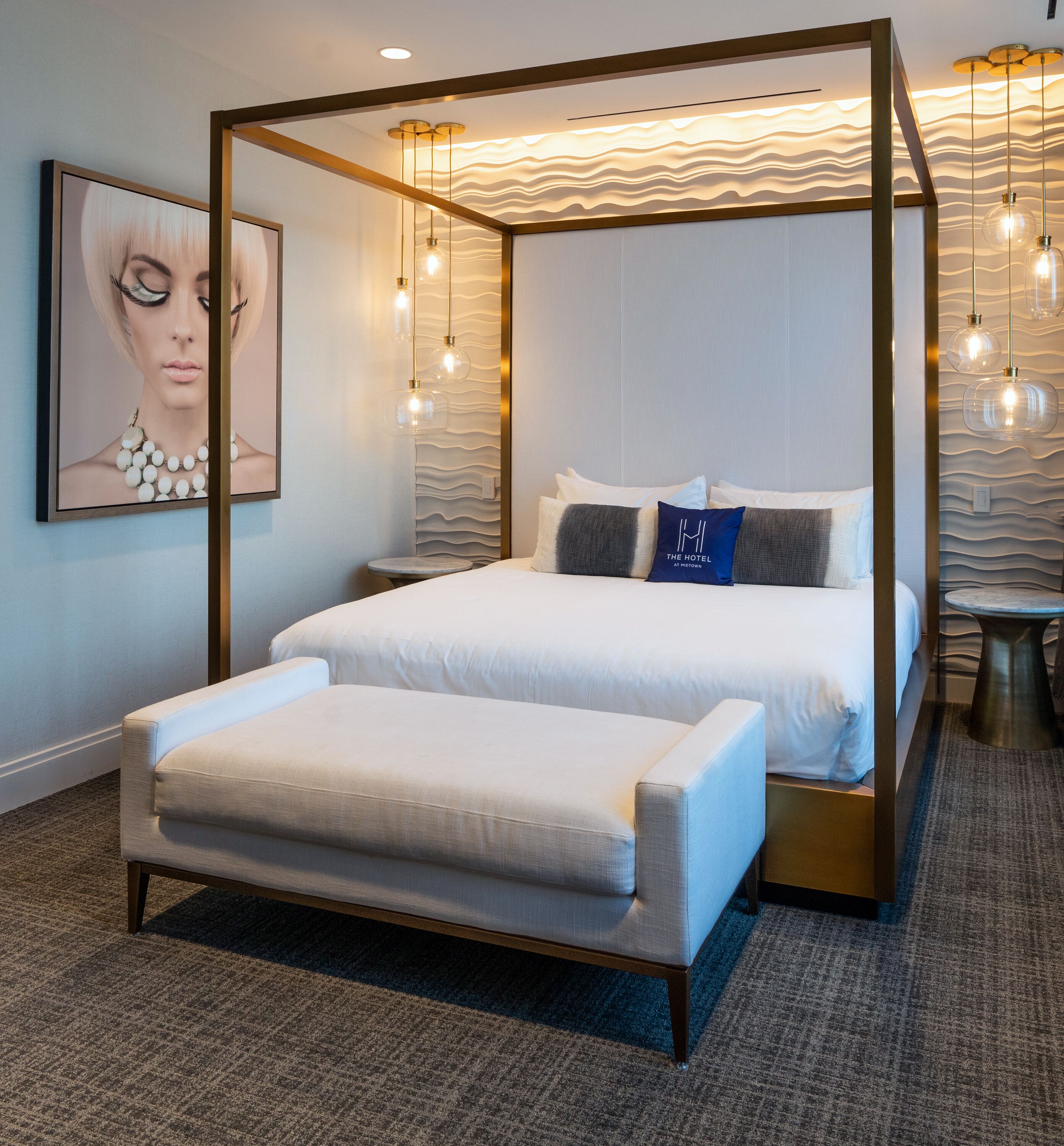 Una cama con dosel, con cubrecama y almohadas blancas, una pequeña mesa redonda y un banco en una habitación de hotel moderna con pared texturizada e iluminación colgante.