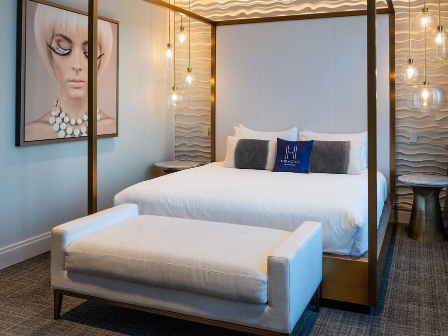 Una cama con dosel, con cubrecama y almohadas blancas, una pequeña mesa redonda y un banco en una habitación de hotel moderna con pared texturizada e iluminación colgante.