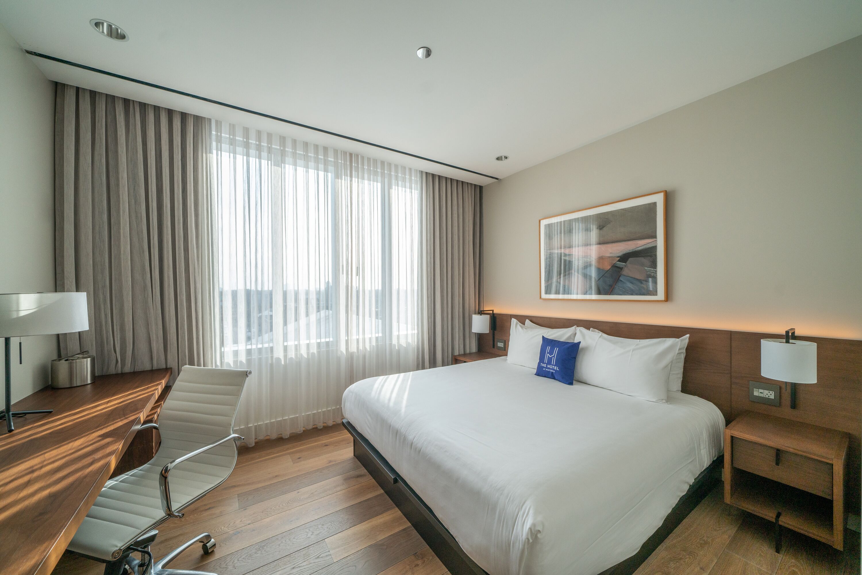 Une chambre d’hôtel avec un grand lit, un bureau avec une chaise, une table de chevet et une fenêtre avec des rideaux.