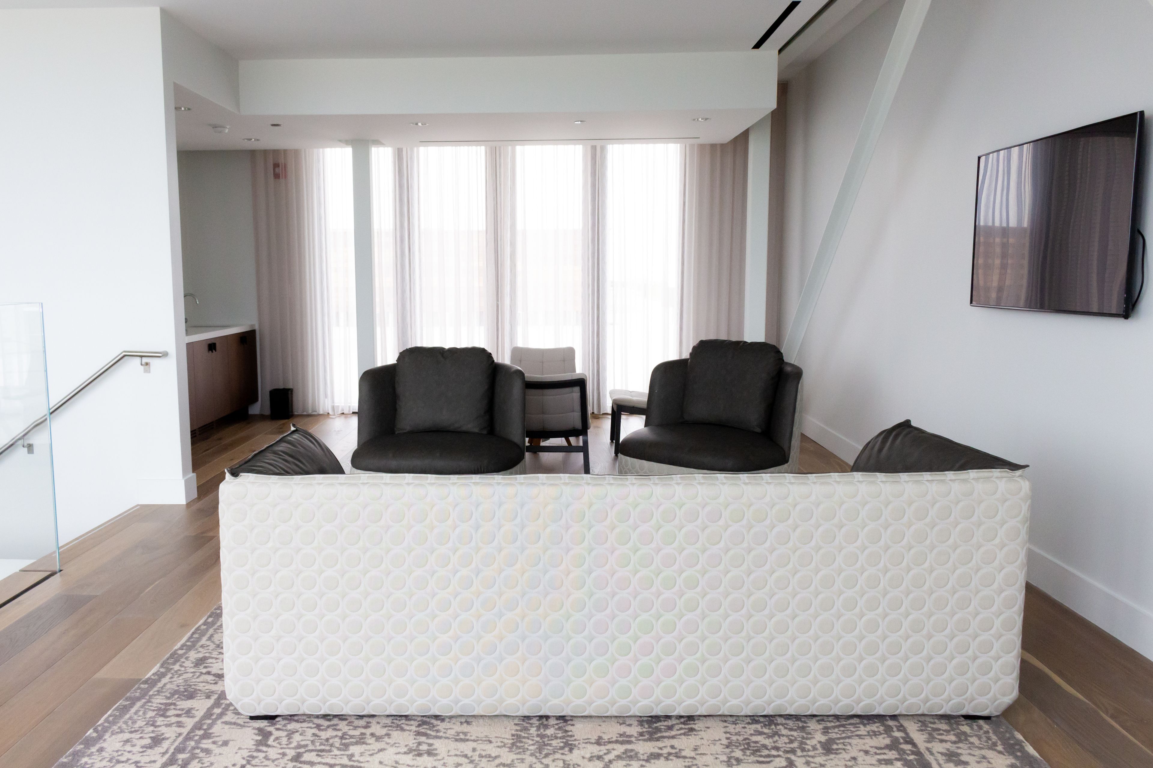 A modern living room with a white sofa, two armchairs, a glass railing, and a large window with curtains.