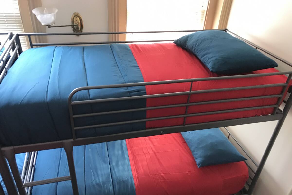 Una cama litera con ropa de cama azul y roja, una escalera metálica y una lámpara de pared.