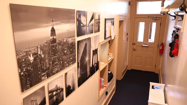Un pasillo con fotografías en blanco y negro de un paisaje urbano, una puerta con ventanal y una pequeña mesa blanca con una silla.
