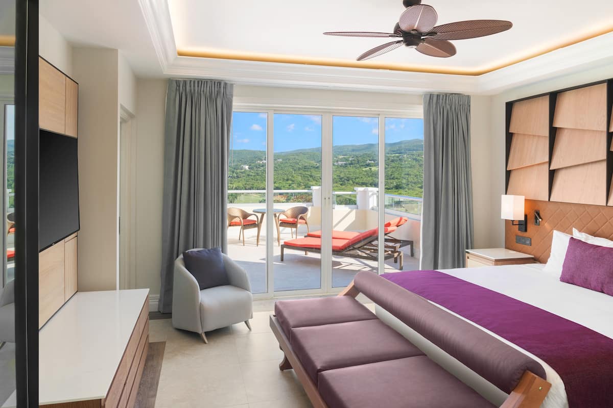 A hotel room with a bed, a sofa, a TV, a ceiling fan, and a balcony with outdoor seating.