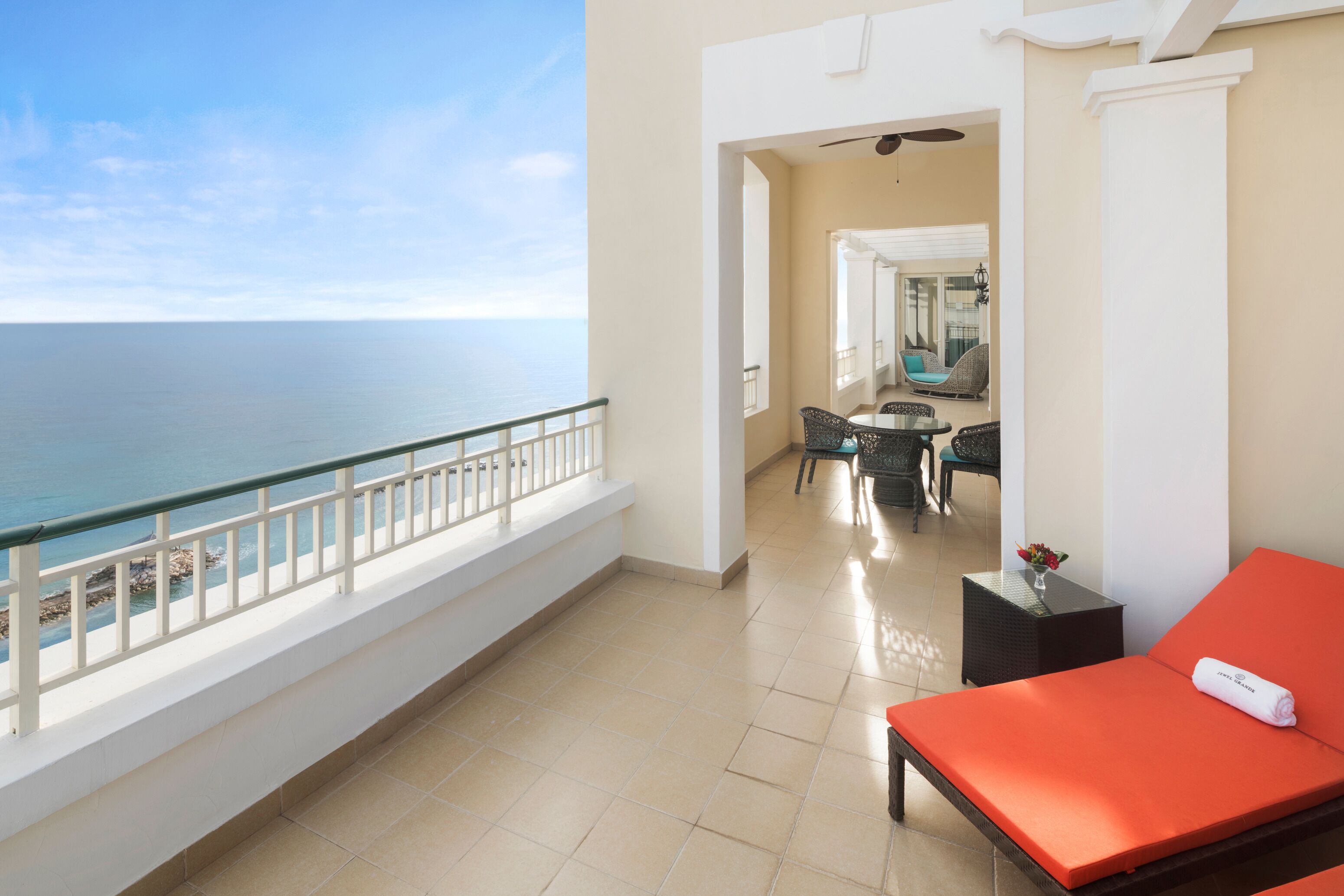 A balcony with a view of the sea, a red lounge chair, and a small table with a vase.