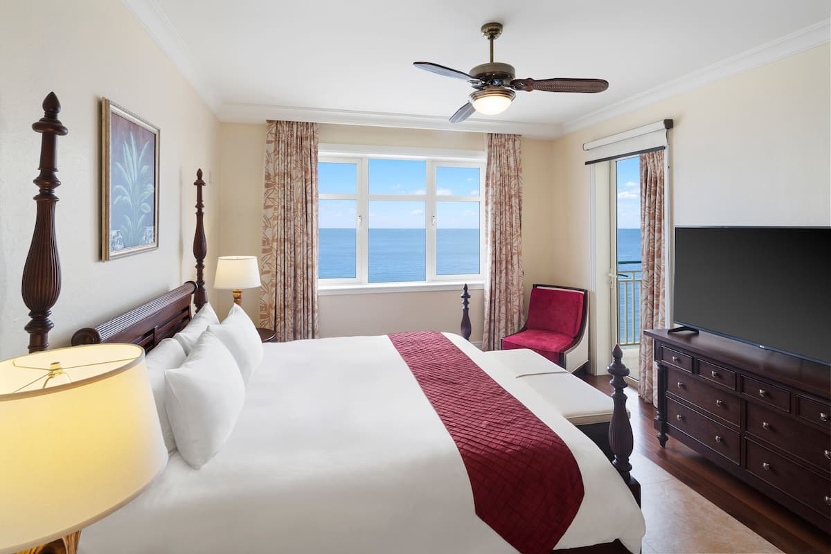 A hotel room with a large bed, a ceiling fan, a TV, a window with a view of the sea, and a red chair.