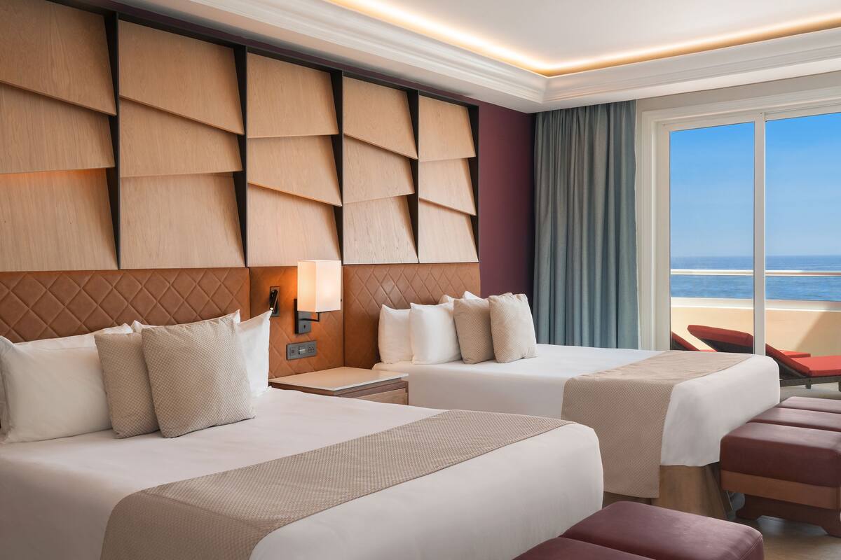 A hotel room with two beds, a large window with a view of the sea, and a wall with a geometric pattern.