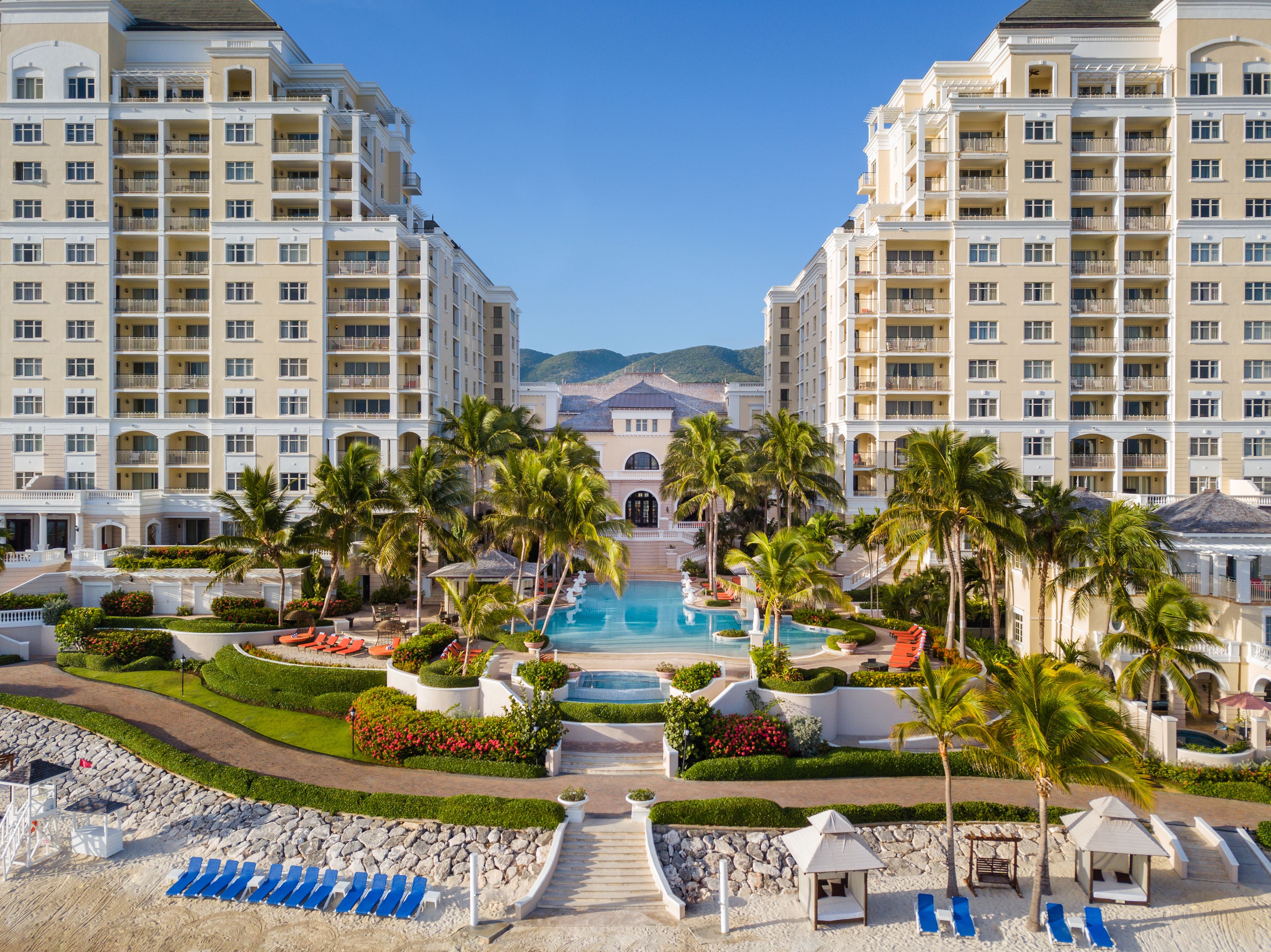 A resort with a central pool, palm trees, and a beach area with lounge chairs.
