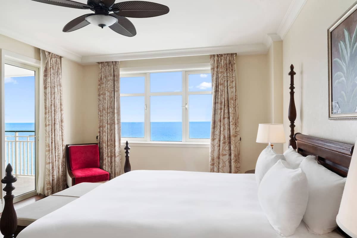 A hotel room with a large bed, a red chair, a ceiling fan, a window with curtains, and a view of the sea.