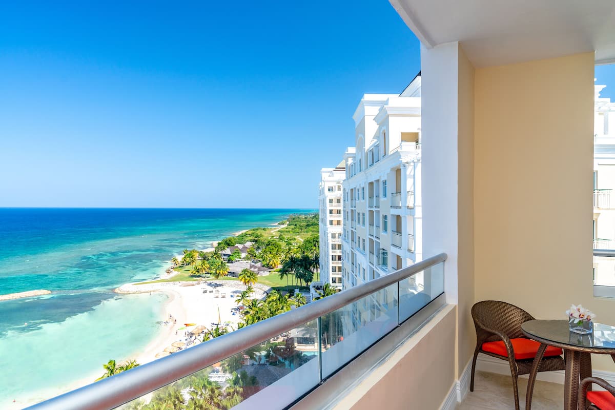A balcony with a view of a beach and ocean, a wicker chair with a cushion, and a small table with a vase.