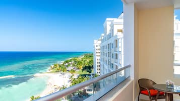 A balcony with a view of a beach and ocean, a wicker chair with a cushion, and a small table with a vase of flowers.