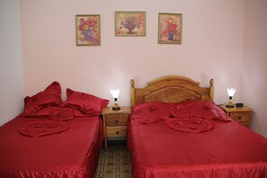 Two single beds with red bedding and decorative pillows, flanked by nightstands with table lamps and framed floral paintings on the wall.