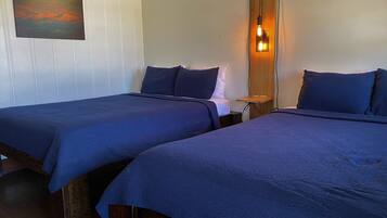 A room with two beds, each with a dark blue bedspread, a wooden headboard, and a nightstand with a lamp. A painting is visible on the wall above the beds.