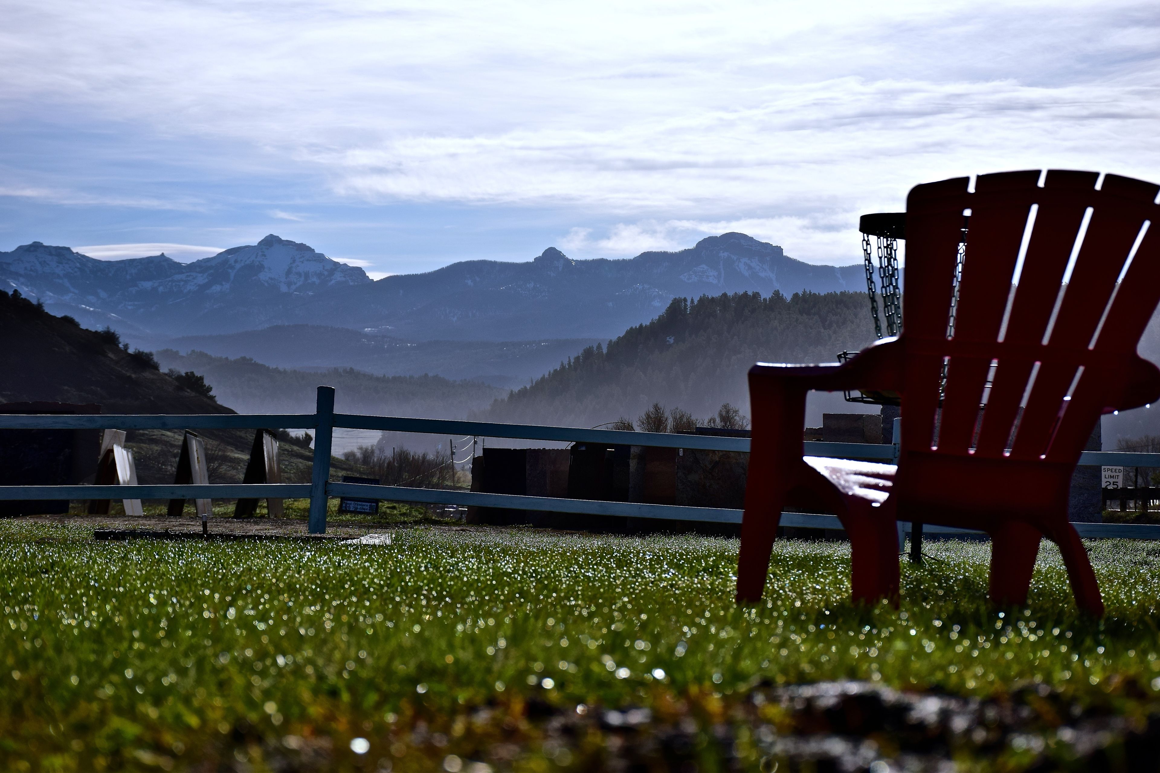 A red Adirondack chair in a grassy area with a view of mountains and a fence in the foreground.