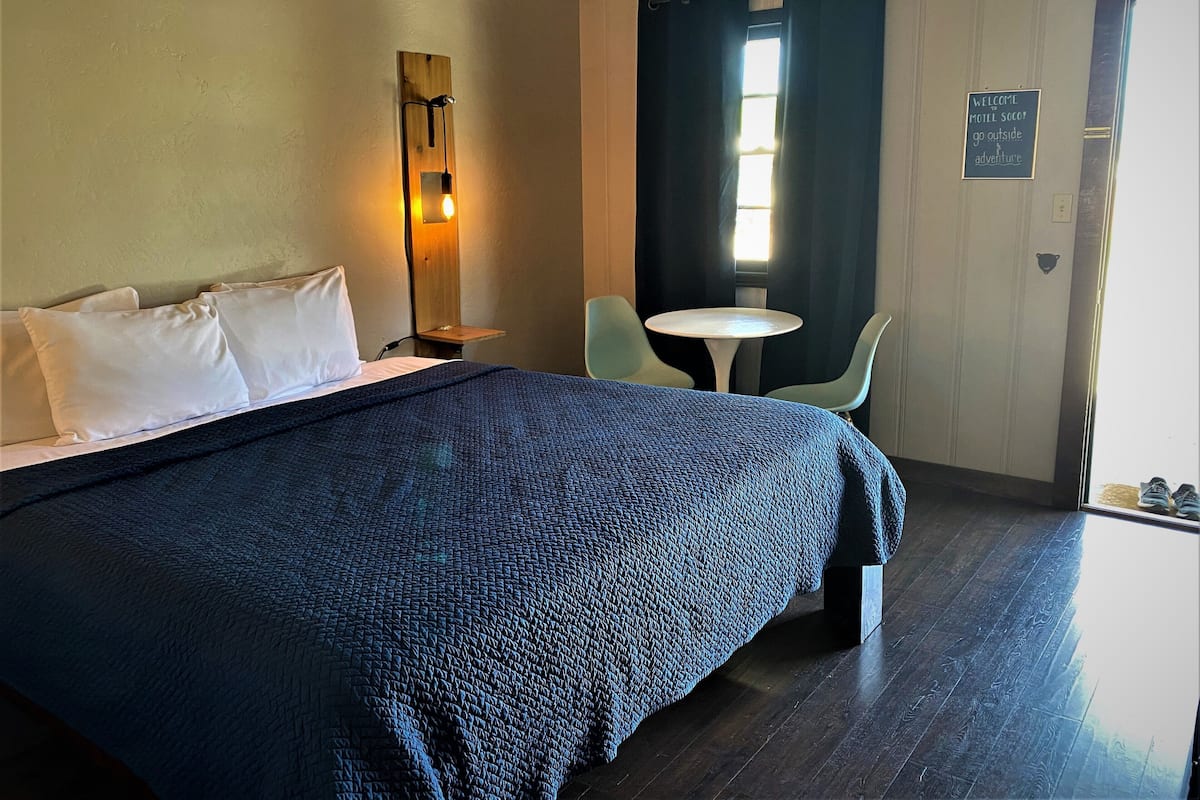A hotel room with a large bed, a bedside table, a small table with chairs, a window with curtains, and a sign on the wall.