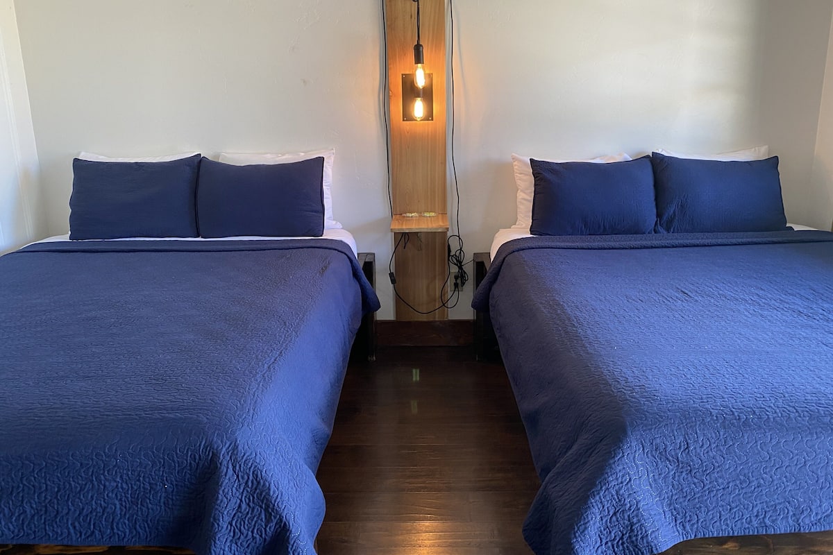 A room with two beds, each with a blue bedspread and pillows. There is a wall-mounted light fixture between the beds.