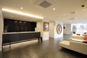 A modern hotel reception area with a dark reception desk, a white sofa, and a large window providing natural light. - Hotel Nanvan Hamanako (Kosai)