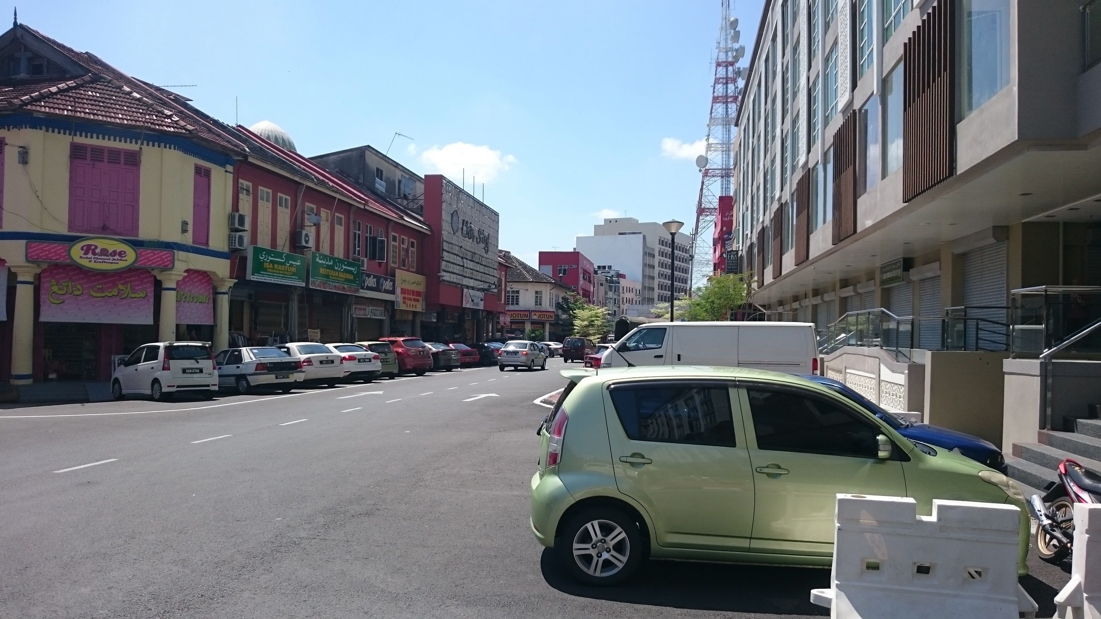Une vue d’une rue bordée de divers commerces, notamment un restaurant et une pharmacie. Plusieurs voitures sont garées le long de la rue, et un fourgon blanc est également visible.