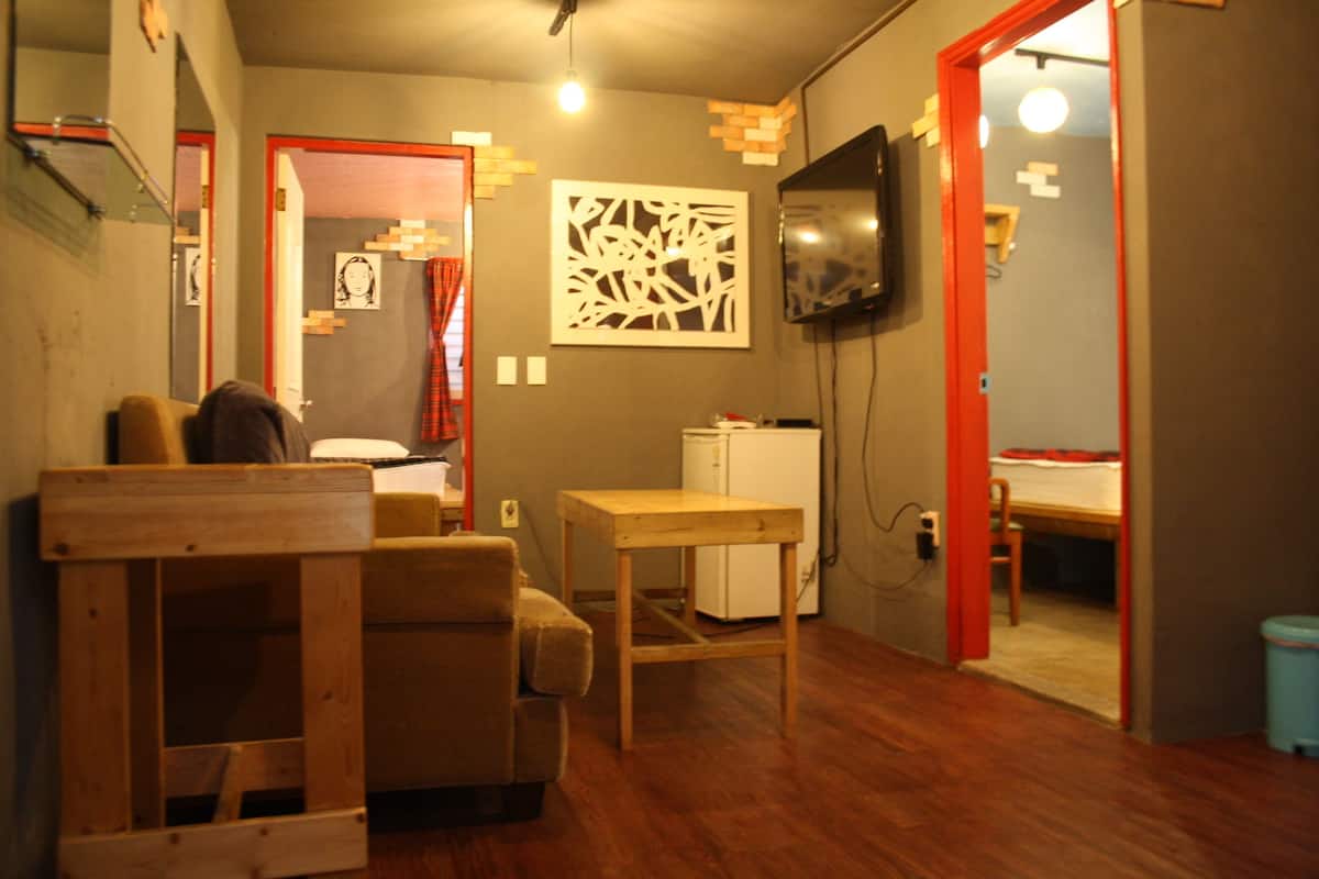 A compact living space with a sofa, a small wooden table, a TV mounted on the wall, and a kitchenette area.