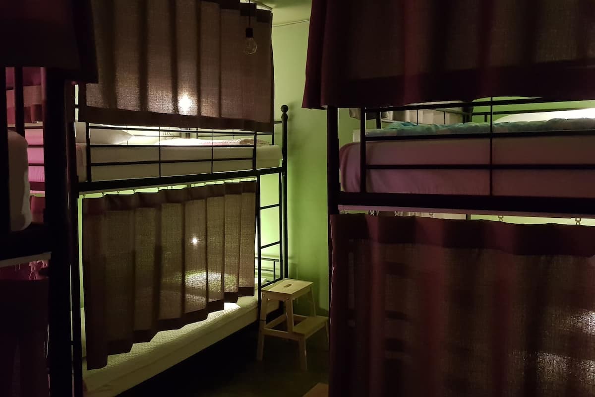 A dormitory room with bunk beds, a small wooden stool, and a window with curtains.