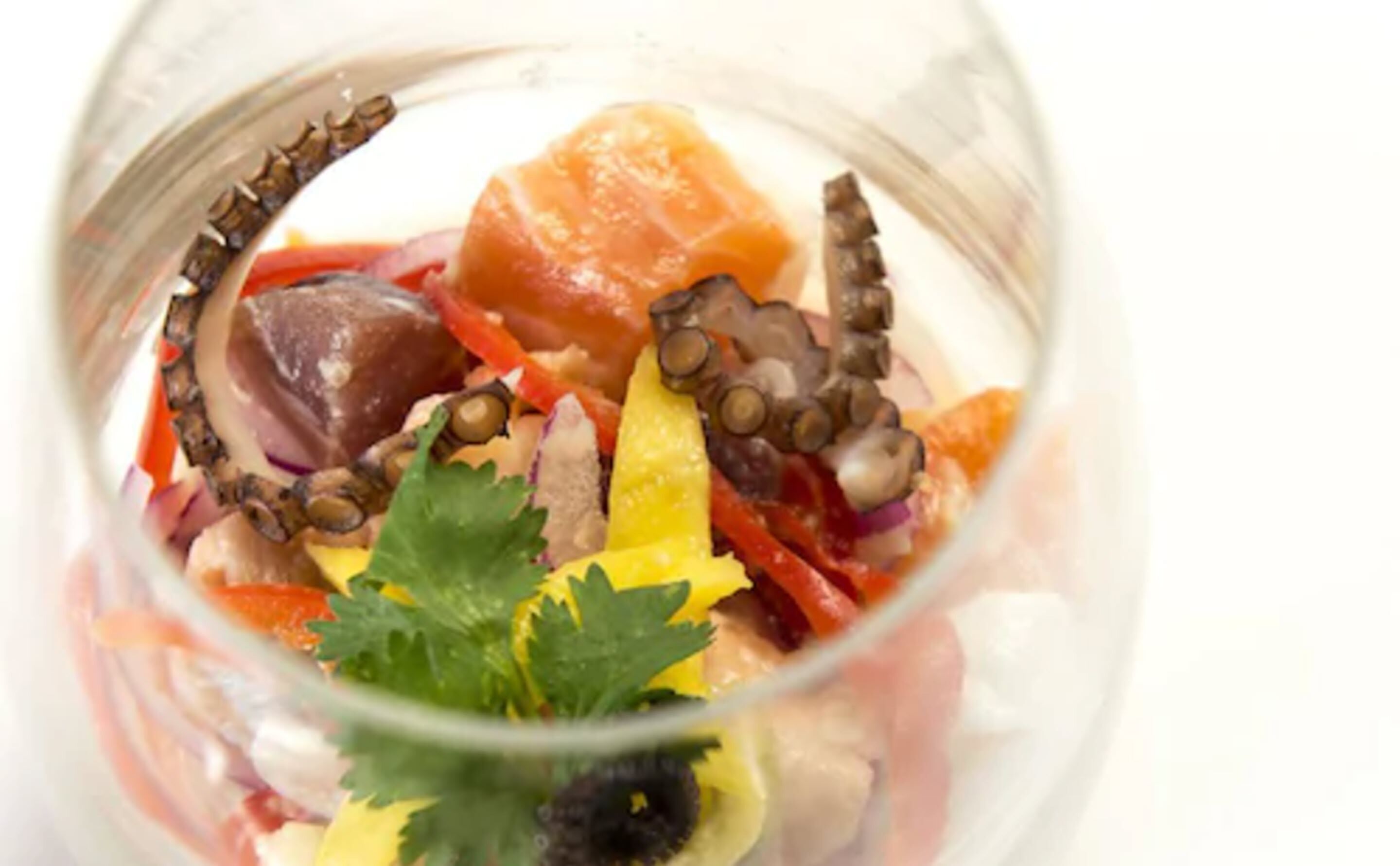 A clear glass bowl containing a seafood salad with octopus, red bell peppers, and a garnish of parsley.