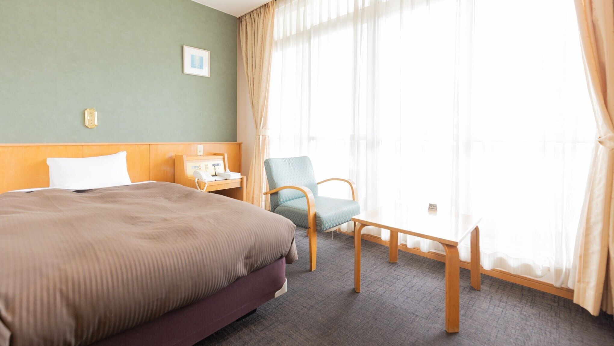 A hotel room with a bed, a chair, a bedside table with a phone, and a window with curtains.