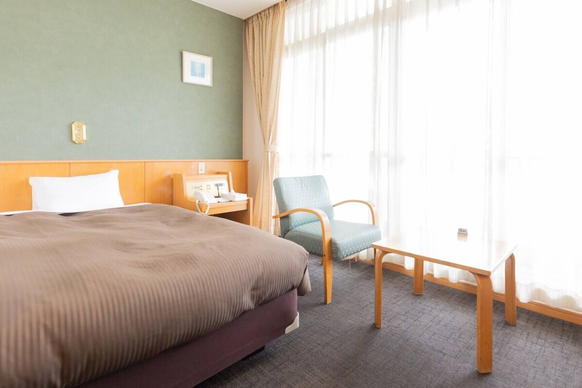 Habitación de hotel con cama, una silla, mesita de noche con teléfono y ventana con cortinas.