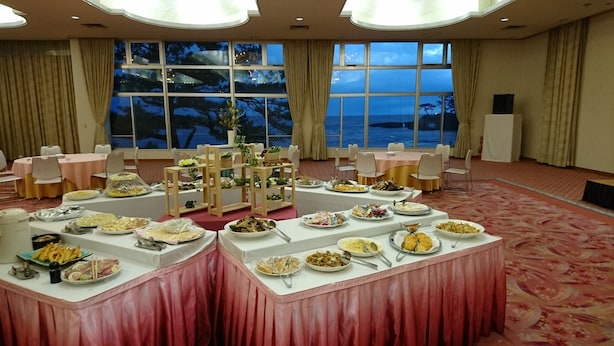 A banquet hall with round tables, a buffet setup, and a view of the sea through large windows. - Hotel Axia Kushikino (Ichikikushikino)