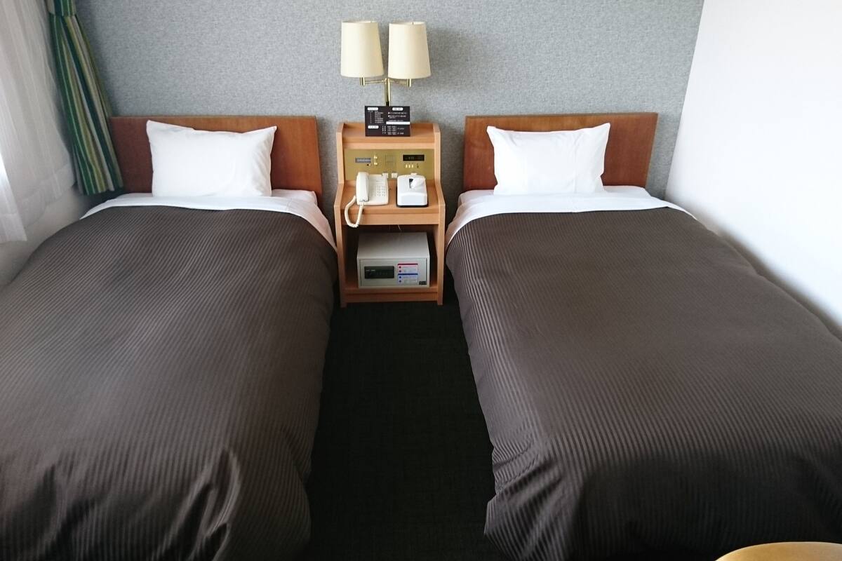 Una habitación de hotel con dos camas individuales, una mesita de noche con teléfono y un reloj digital, y una lámpara.