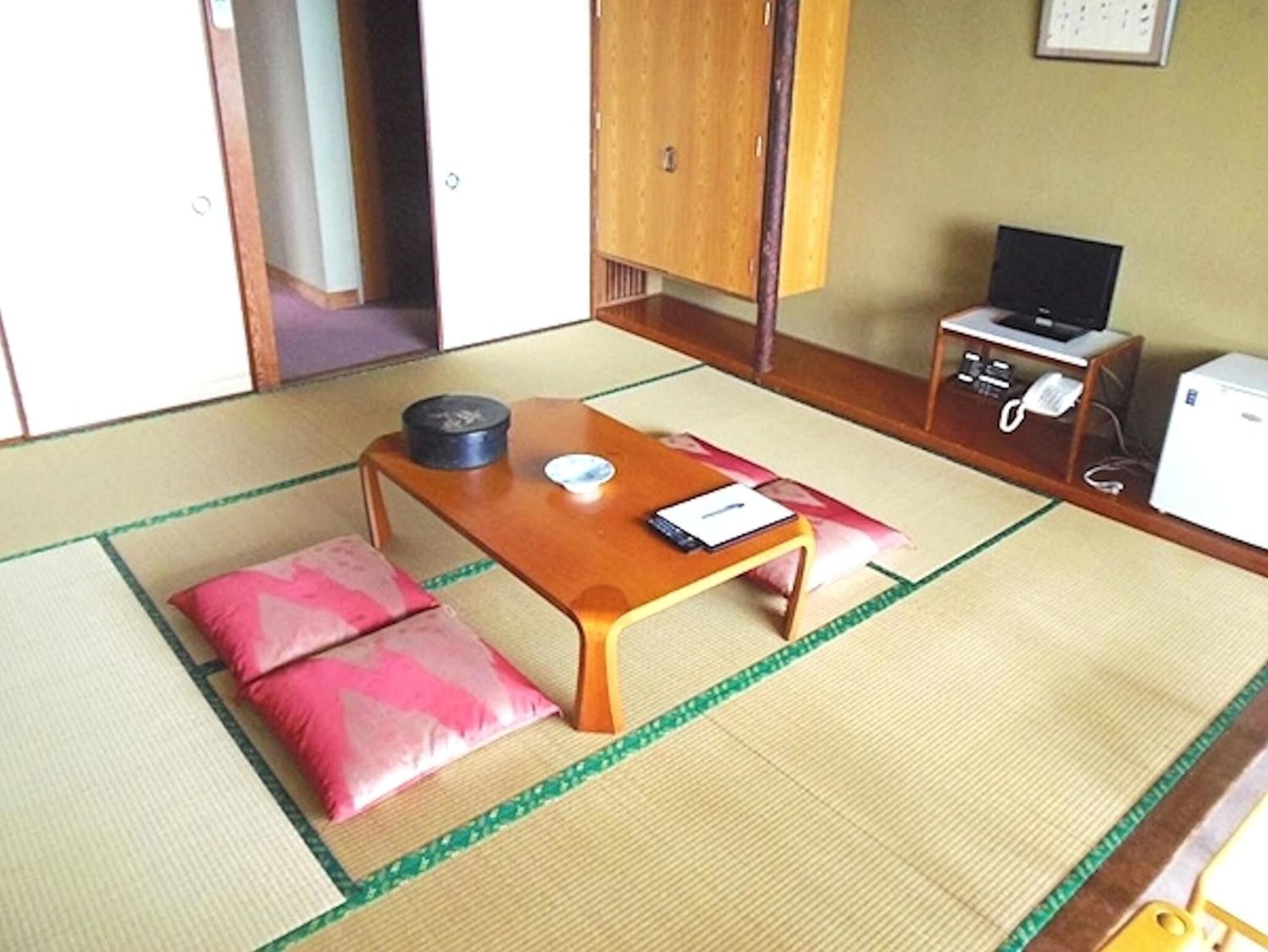 A traditional Japanese room with tatami flooring, a low wooden table, pink cushions, a television, and a small refrigerator.