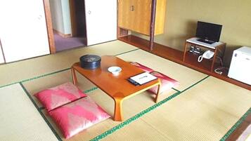 A traditional Japanese room with tatami flooring, a low wooden table, pink cushions, a television, and a small refrigerator.